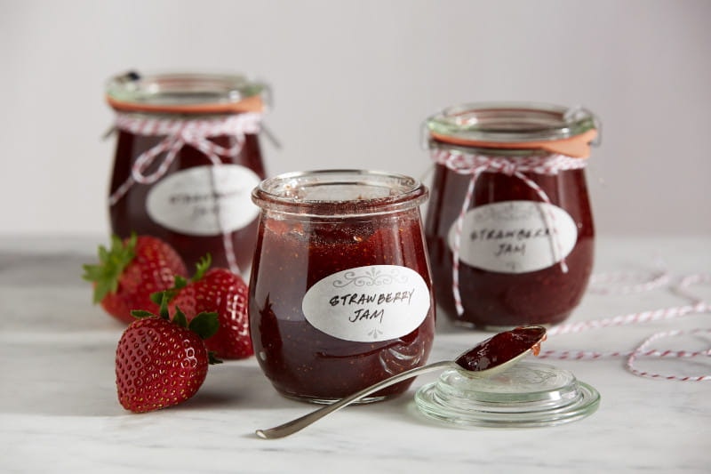 Three jars of small batch strawberry jams sitting next to three strawberries. A thin ribbon is tied around two of the jars.