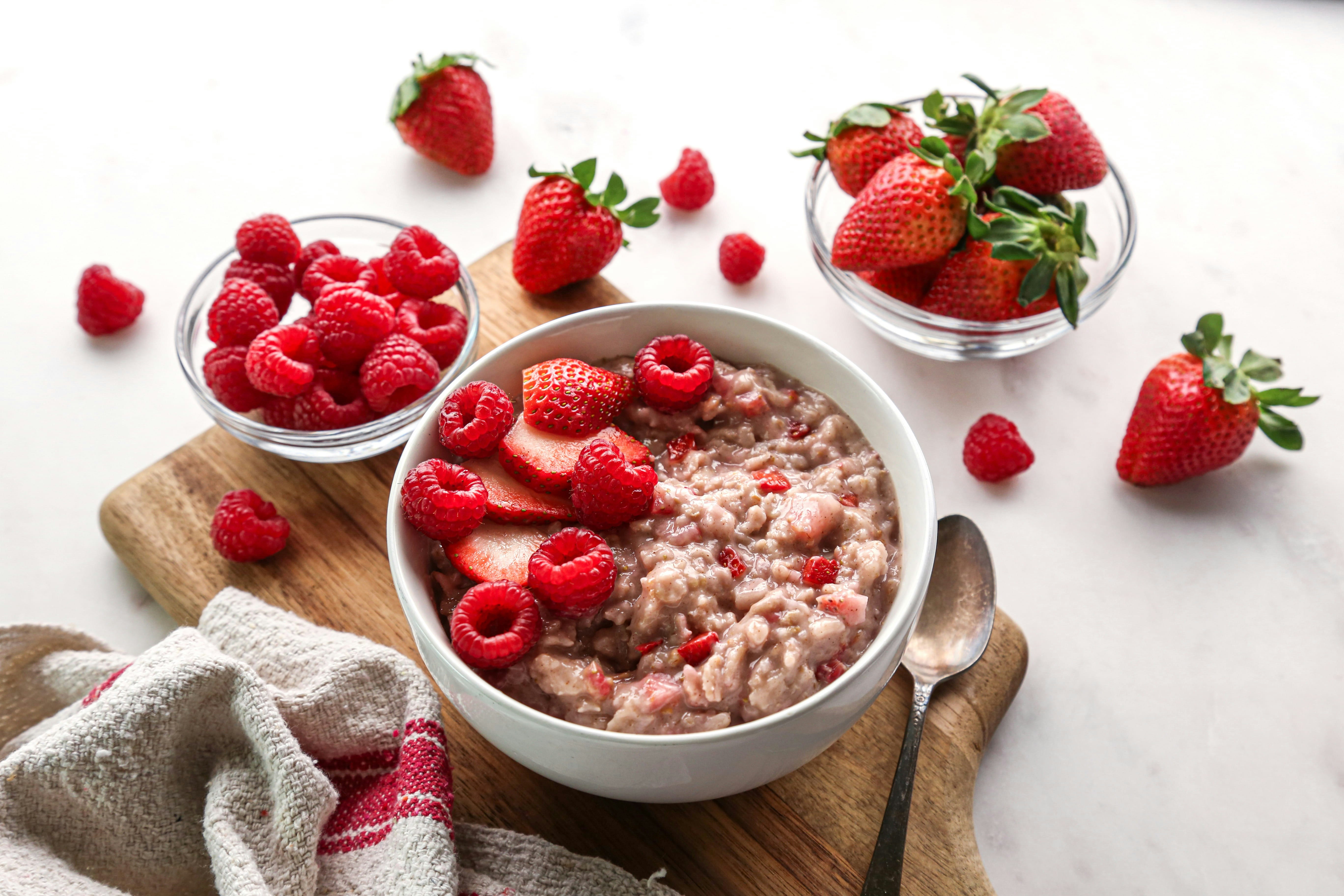 A bowl of strawberry raspberry oatmeal sitting on a cutting board next to two cups of berries.